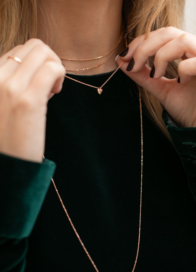 Zarte Halskette aus Roségold mit kleinem Herzanhänger, kombiniert im eleganten Layering-Look am Hals einer blonden Frau getragen.