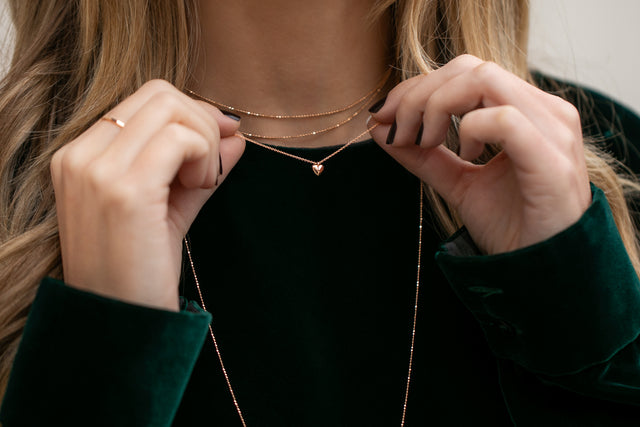 Zarte Halskette aus Roségold mit kleinem Herzanhänger, kombiniert im eleganten Layering-Look am Hals einer blonden Frau getragen.