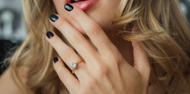  blonde Frau mit Verlobungsring aus Roségold mit weißem Diamanten streicht sich mit Finger über Lippe 