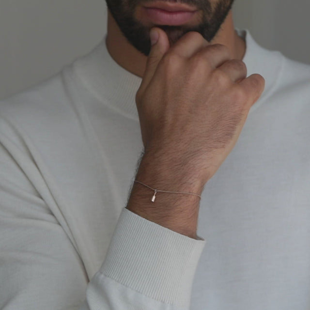 Ein schlichtes Stoffarmband mit einem Anhaenger aus Roségold. Der Anhaenger stellt 1 Gram Liebe dar und ist geformt wie ein kleines Gramm-Gewicht. Getragen an einem Maennerhandgelenk.